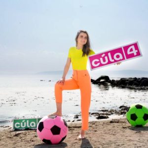 Photo of Cúla4 host Niamh Ní Chróinín stood on a beach holding a sign saying 'Cúla4;